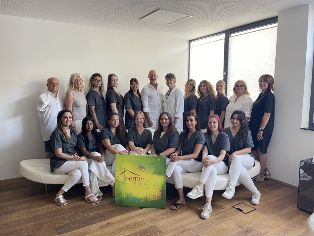 Das Praxisteam aus 21 Personen, 8 Personen sitzen auf einem Sofa, die anderen stehen dahinter, alle in dunkelblauer und weißer Praxis-Kleidung.